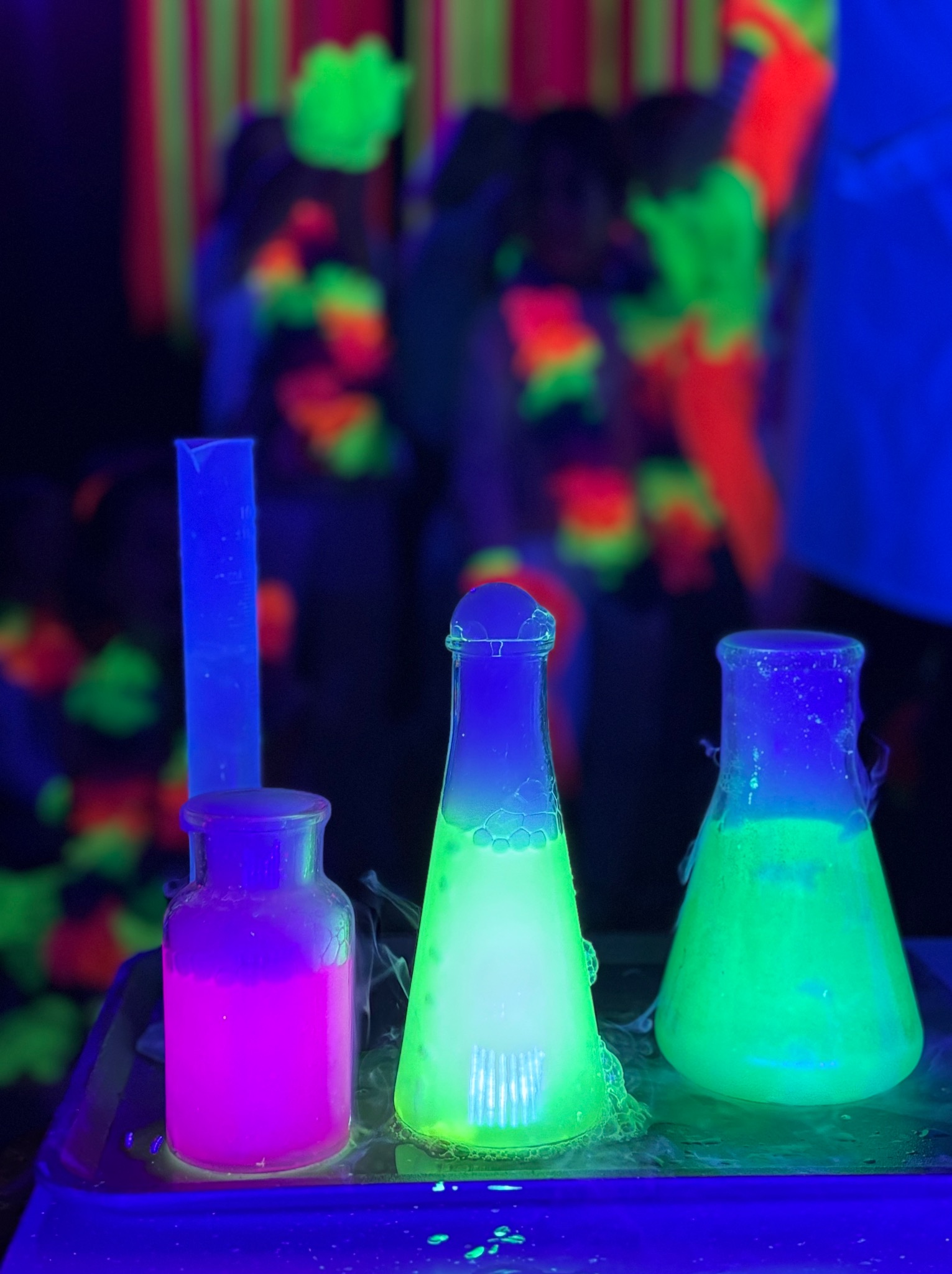 Eksperymenty chemiczne z użyciem fluorescencyjnych płynów w kolbach i probówkach, podświetlone światłem UV, w tle sylwetki osób w neonowych dekoracjach.