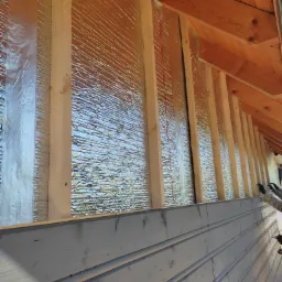 Ocieplanie ściany od wewnątrz folią termoizolacyjną i drewnianą konstrukcją nośną, widoczne deski elewacyjne pomalowane na szaro w dolnej części ściany.