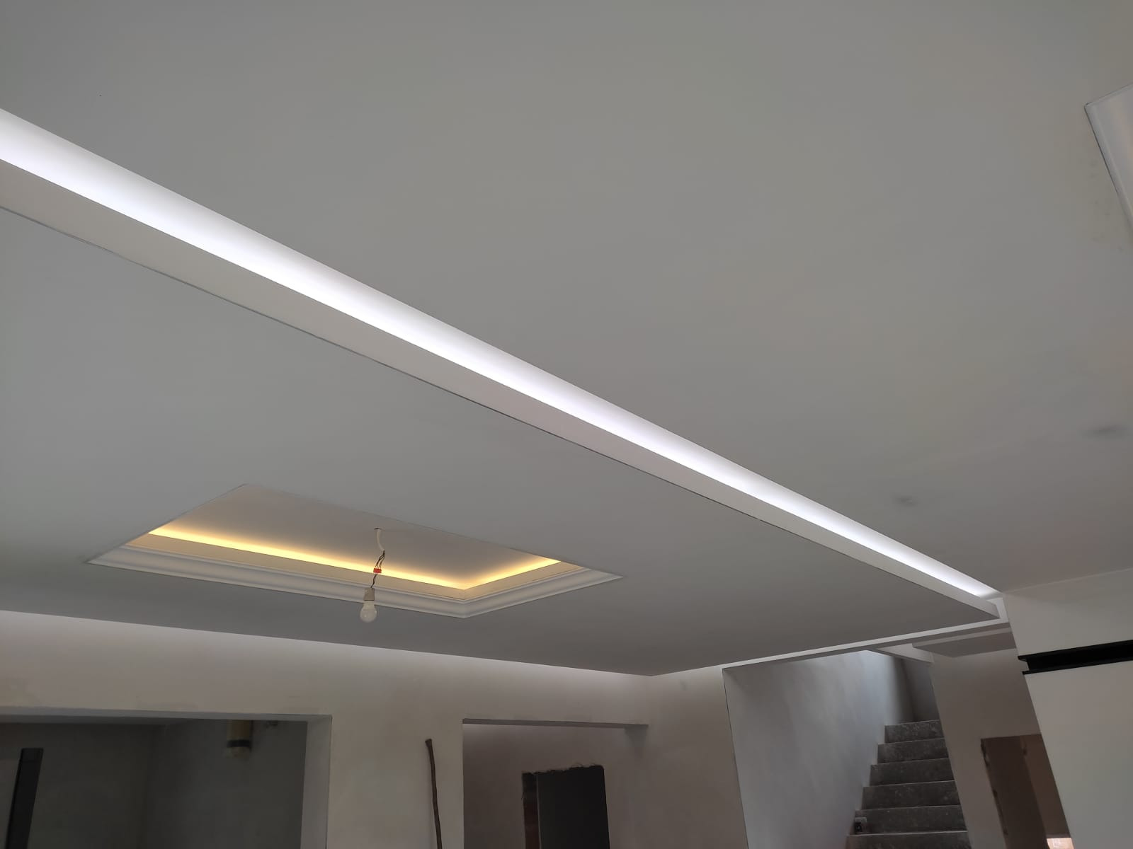 Wykończony sufit z podwieszanym oświetleniem LED i dekoracyjnym wgłębieniem z dodatkowym oświetleniem, widok na schody w tle.