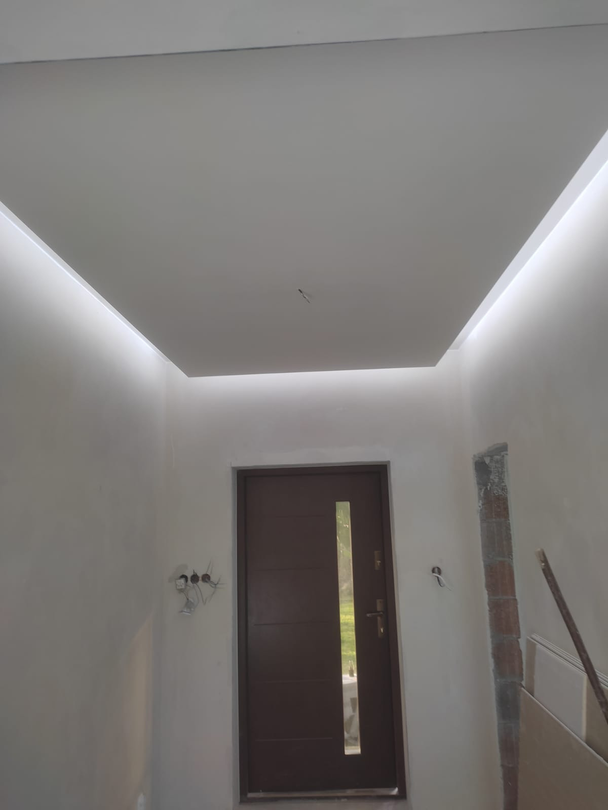 Wykończone wnętrze z podwieszonym sufitem z oświetleniem LED wzdłuż krawędzi, brązowe drzwi wejściowe z długim przeszkleniem, ściany przygotowane do malowania, widoczne przewody elektryczne...