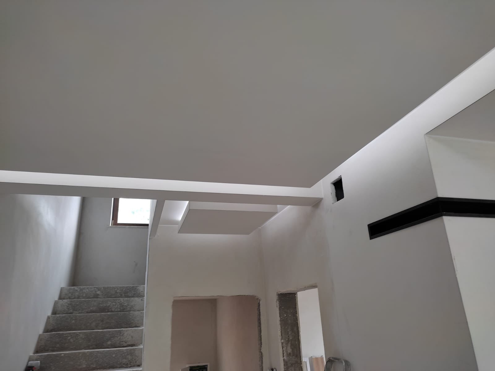 Wnętrze w trakcie remontu: surowe schody, białe ściany z widocznymi instalacjami i elementami dekoracyjnymi pod sufitem, okno w tle.