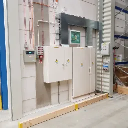 Rozbudowana instalacja elektryczna w hali magazynowej, widoczne szafy elektryczne, przewody w korytkach, zabezpieczenia i elementy uziemiające, z żółtymi oznaczeniami bezpieczeństwa.