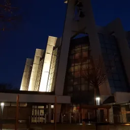Nowoczesny kościół oświetlony nocą, z widoczną wieżą z dzwonami i szklaną fasadą, oświetlony lampami ulicznymi.