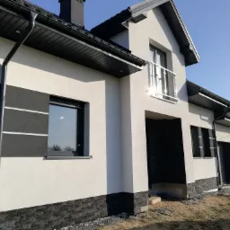 Nowoczesny dom z białą elewacją, czarnymi akcentami w postaci okładziny imitującej kamień i grafitową stolarką okienną, widoczne orynnowanie i fragment balkonu z metalową balustradą.