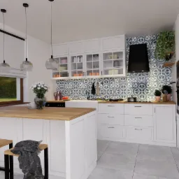 Jasna kuchnia z wyspą, białymi szafkami, drewnianym blatem, wzorzystą ścianą nad blatem, wiszącymi lampami i kwiatami w wazonie.