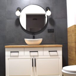 Vertical - Nowoczesna łazienka z ciemnoszarą ścianą z płytek, białą szafką pod umywalkę z drewnianym blatem, okrągłym lustrem z lampami kulami i czarną armaturą.