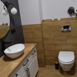 Vertical - Nowoczesna łazienka z drewnianymi akcentami: blat z jasnego drewna, biała umywalka nablatowa, okrągłe lustro z oświetleniem, toaleta wisząca i dekoracja ścienna w kształcie owcy z rolek papieru...