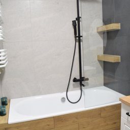 Vertical - Biała wanna z drewnianą obudową i czarną armaturą prysznicową na tle szaro-białych płytek, uzupełniona o drewniane półki i grzejnik drabinkowy.