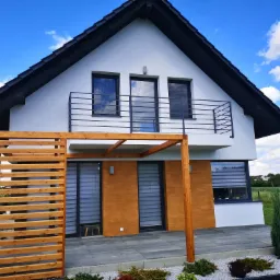 Nowoczesny dom jednorodzinny z drewnianą pergolą i tarasem wyłożonym szarą kostką, ozdobiony białym żwirem i niskimi krzewami ozdobnymi, na tle błękitnego nieba z chmurami.