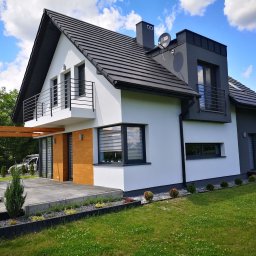 Vertical - Nowoczesny dom jednorodzinny z białymi ścianami, grafitowym dachem i elementami drewnianymi, taras z pergolą i starannie zagospodarowany ogród z niskimi krzewami i trawą.