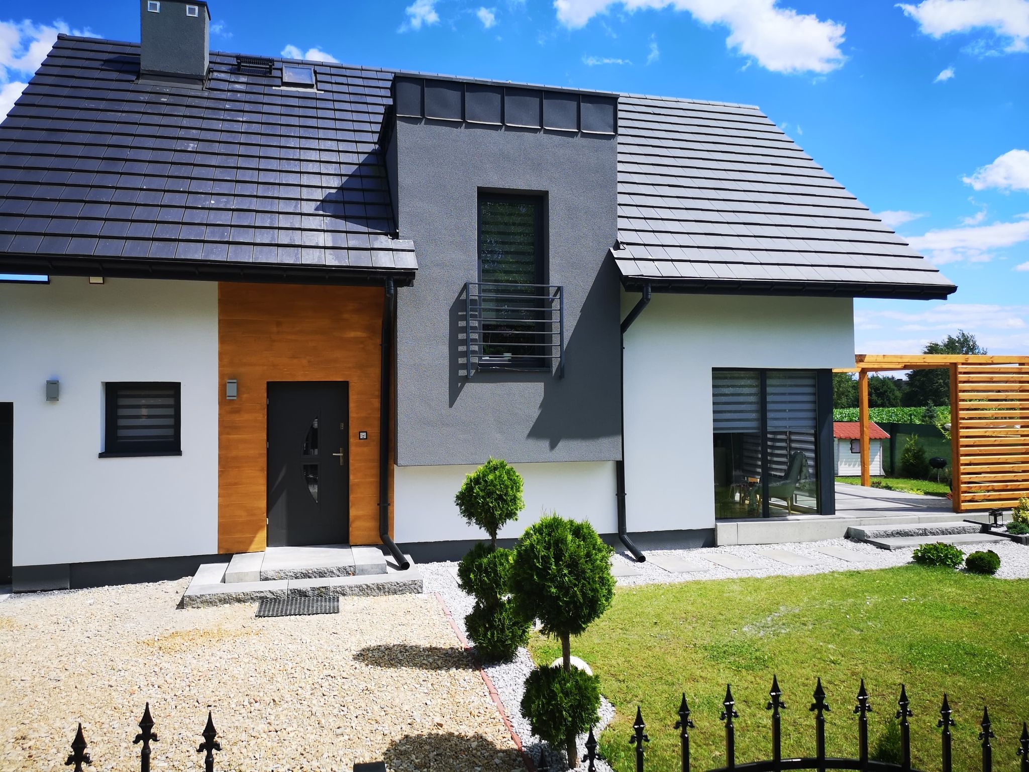 Nowoczesny dom jednorodzinny z grafitową stolarką okienną i drewnianą elewacją, zagospodarowany teren z ozdobnymi krzewami i żwirową ścieżką.