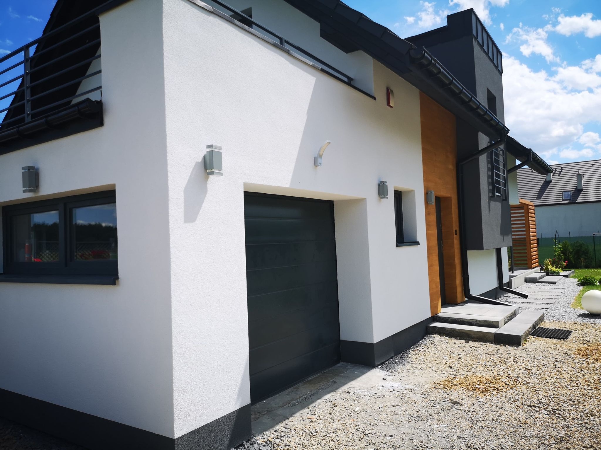 Nowoczesny budynek z garażem i elementami drewna na elewacji, widok z zewnątrz, szara brama garażowa, białe ściany, czarne elementy dachu i wykończenia, teren wokół budynku wysypany żwirem.