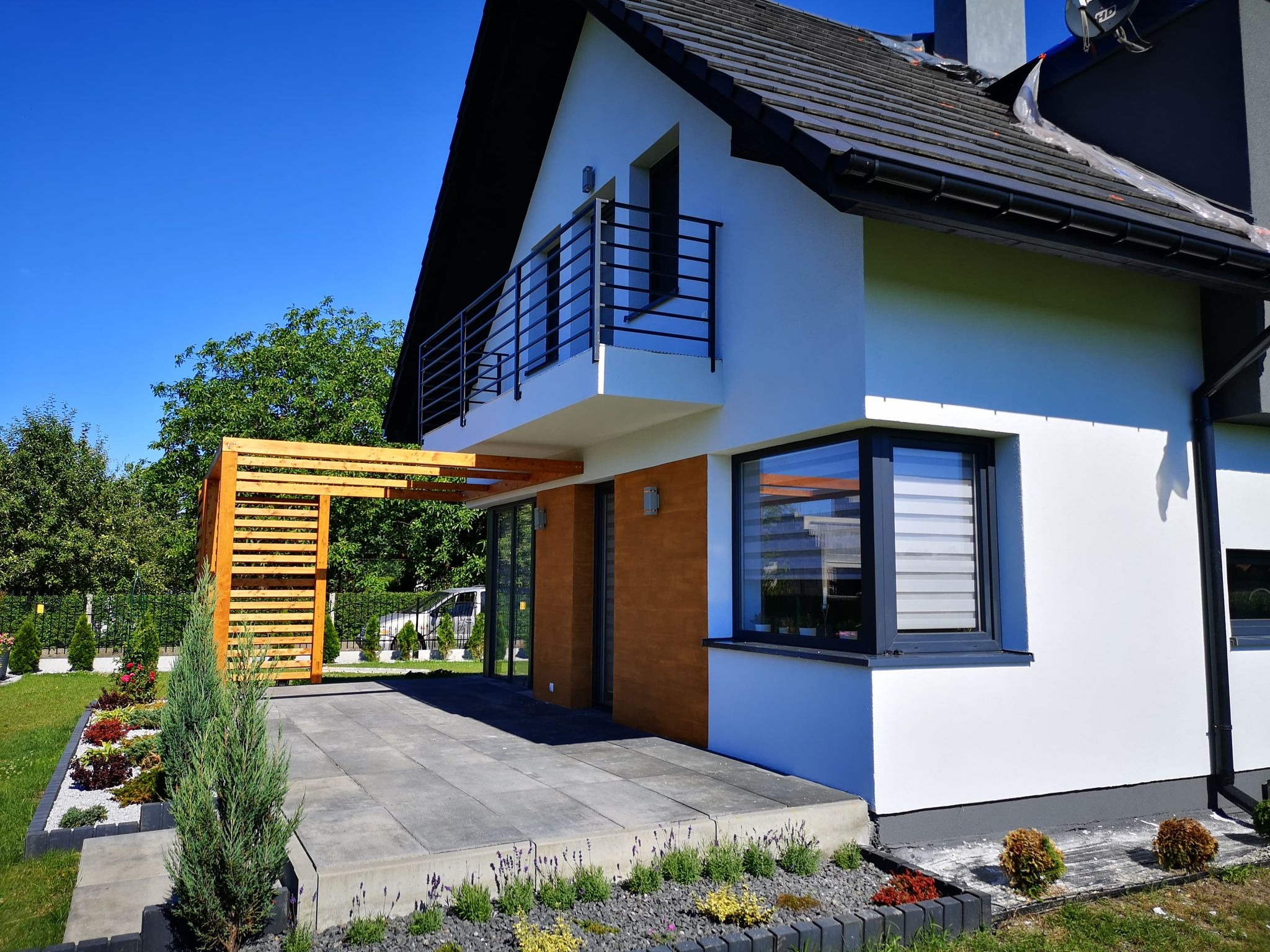 Nowoczesny dom jednorodzinny z tarasem wyłożonym szarą kostką, drewnianą pergolą i zadbanym ogrodem z kolorowymi rabatami.