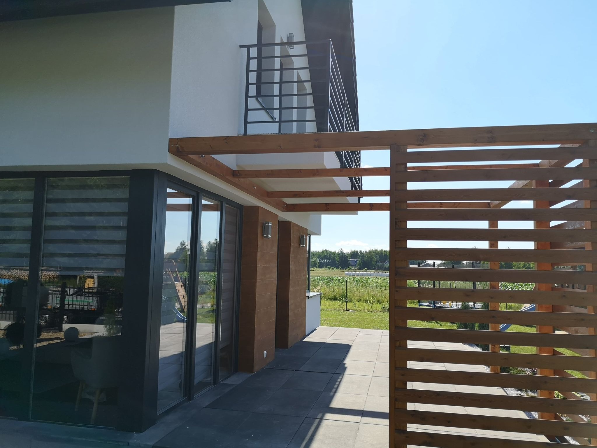 Nowoczesny taras z drewnianą pergolą i przesuwnymi szklanymi drzwiami, widok na pole i błękitne niebo.