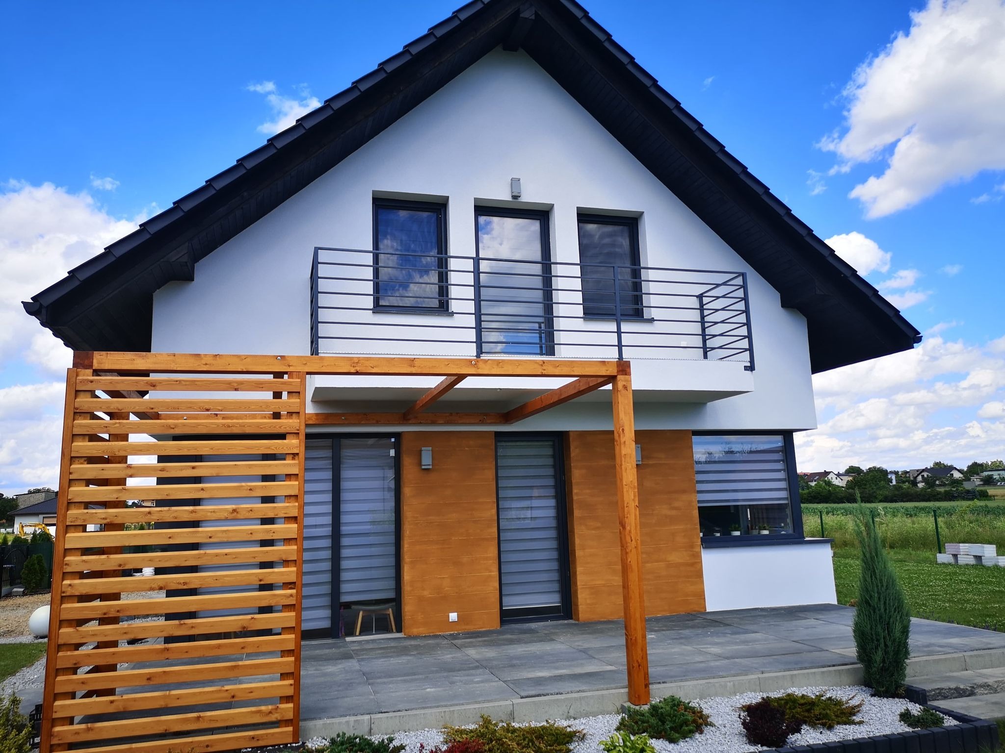 Nowoczesny dom jednorodzinny z drewnianą pergolą i tarasem wyłożonym szarą kostką, ozdobiony białym żwirem i niskimi krzewami ozdobnymi, na tle błękitnego nieba z chmurami.