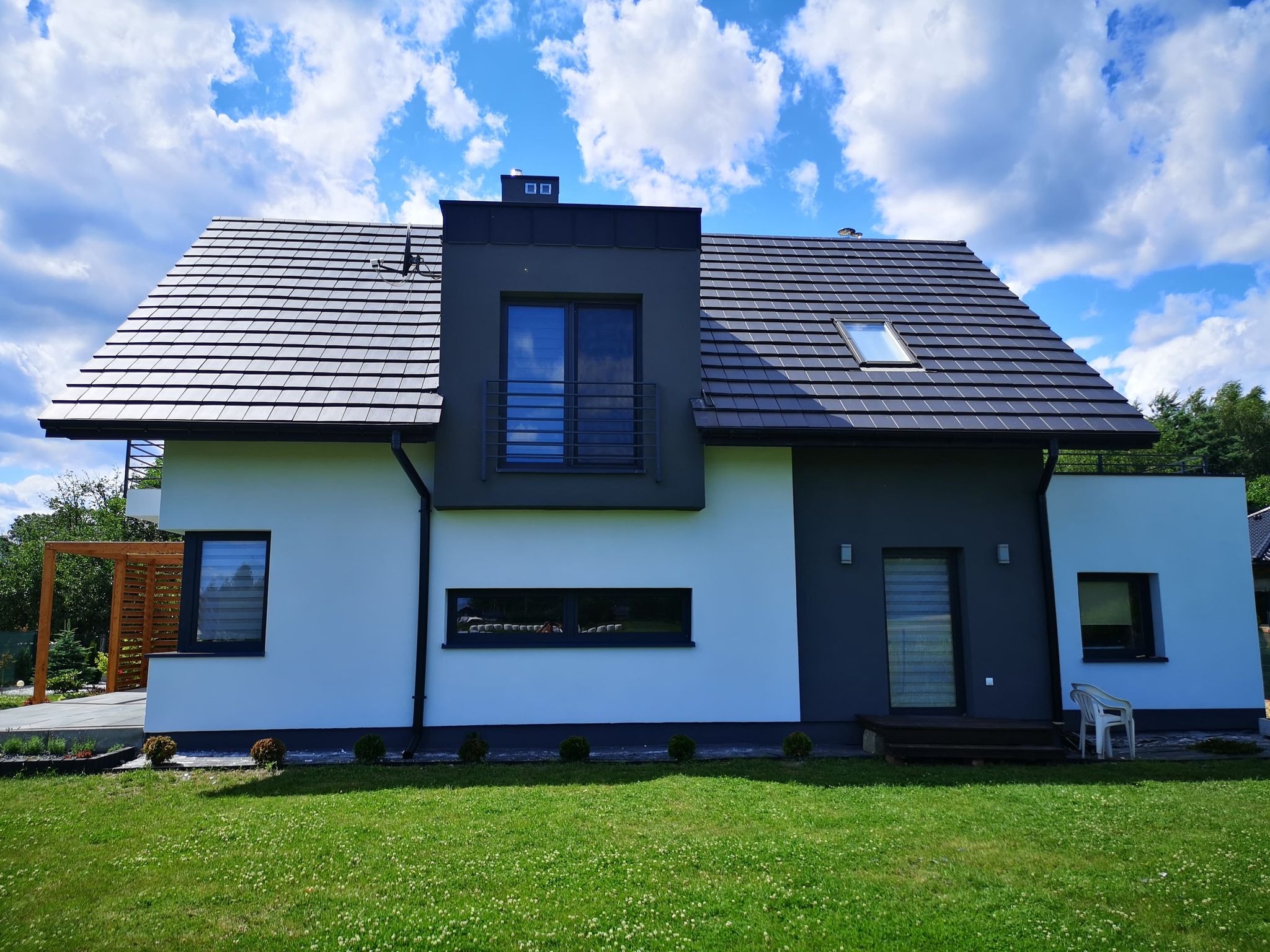 Nowoczesny dom jednorodzinny z dwuspadowym dachem pokrytym ciemną dachówką, z elementami elewacji w odcieniach szarości i bieli, widoczny z perspektywy ogrodu w słoneczny dzień.