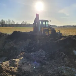 Żółto-czarna koparko-ładowarka Volvo na polu, pracująca przy wykopie ziemnym w słoneczny dzień. W tle widoczne drzewa i błękitne niebo.