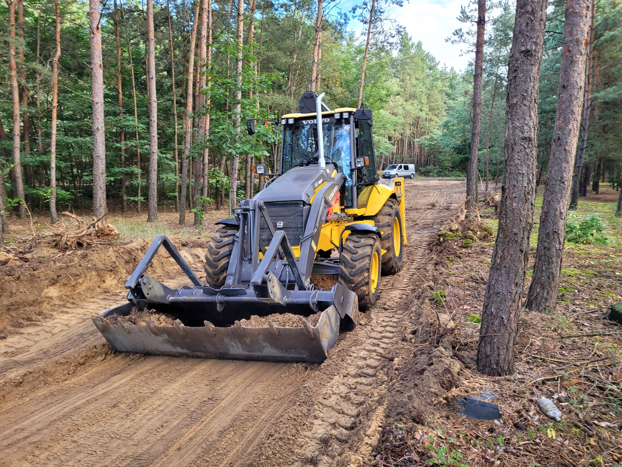 Żółto-szara ładowarka Volvo na piaszczystej drodze w lesie, widoczny ślad opon i wykopana ziemia, w tle biały van.