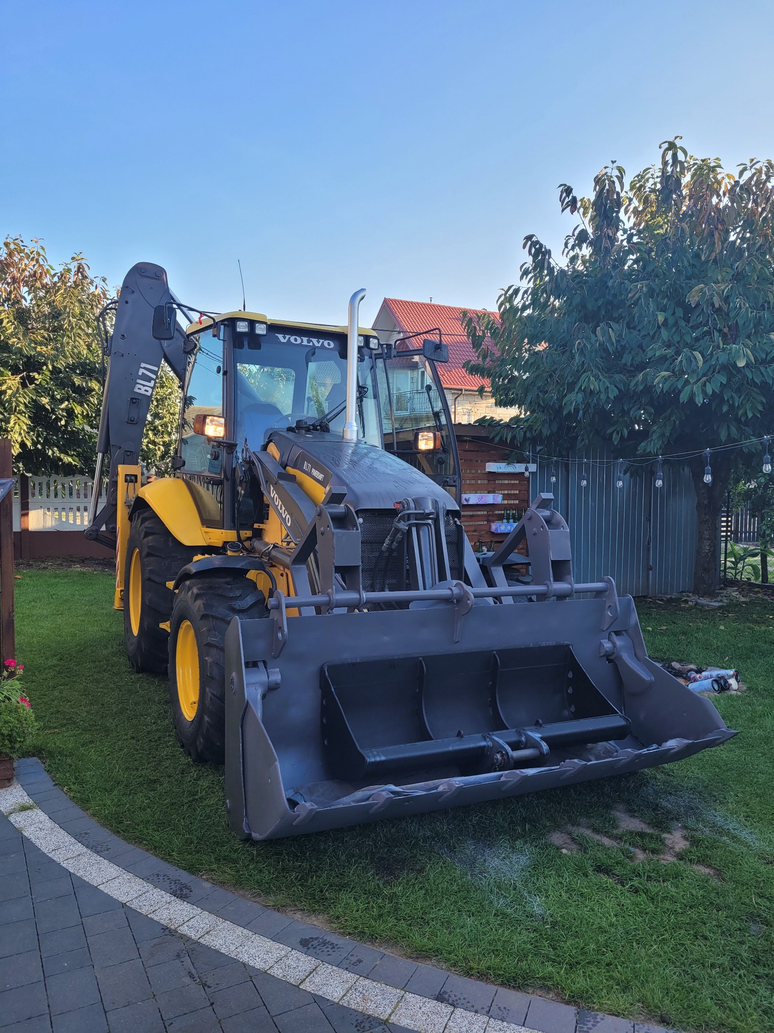 Żółto-szara koparko-ładowarka Volvo BL71 zaparkowana na trawniku przed domem z oświetleniem ogrodowym.