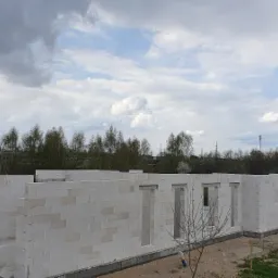 Wznoszenie ścian parterowego domu z białych bloczków, widoczne otwory okienne i drzwiowe, na zewnątrz niebieska toaleta przenośna i zaparkowany niebieski samochód dostawczy, w tle drzewa...