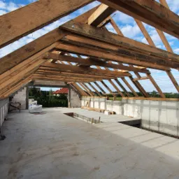 Surowa konstrukcja dachu z drewnianych belek na murowanym domu w trakcie budowy, widok z wnętrza z betonową posadzką i fragmentami ścian z białego betonu kom&oacute;rkowego, błękitne niebo w tle.