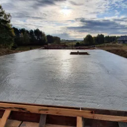 Świeżo wylana płyta fundamentowa pod dom jednorodzinny w otoczeniu zieleni, widoczna drewniana szalówka i zbrojenie w centralnej części, w tle zabudowa jednorodzinna pod błękitnym niebem.