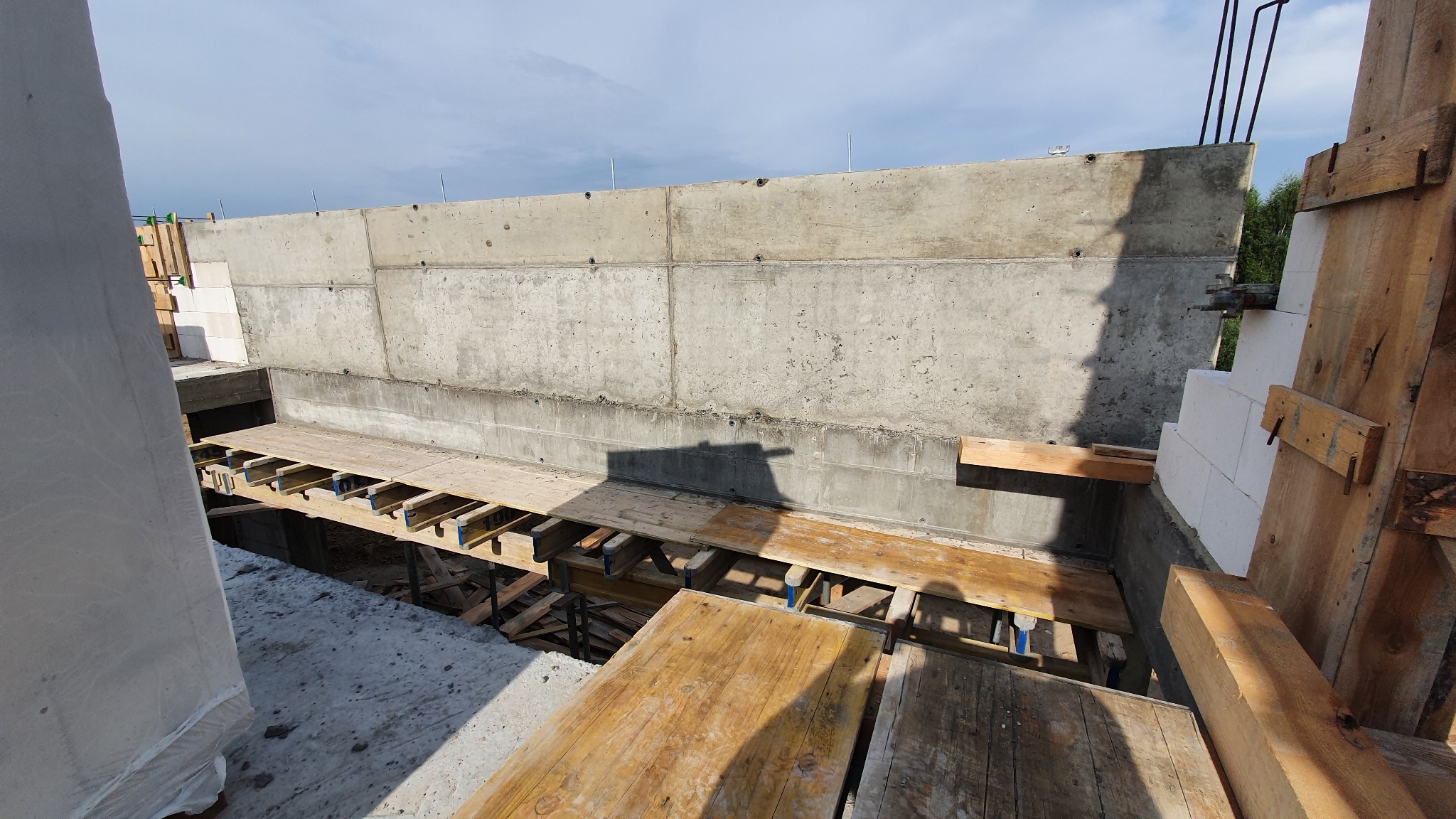 Wysoki betonowy fundament z widocznym deskowaniem i wystającymi prętami zbrojeniowymi, częściowo oszalowany białymi bloczkami, w słoneczny dzień.