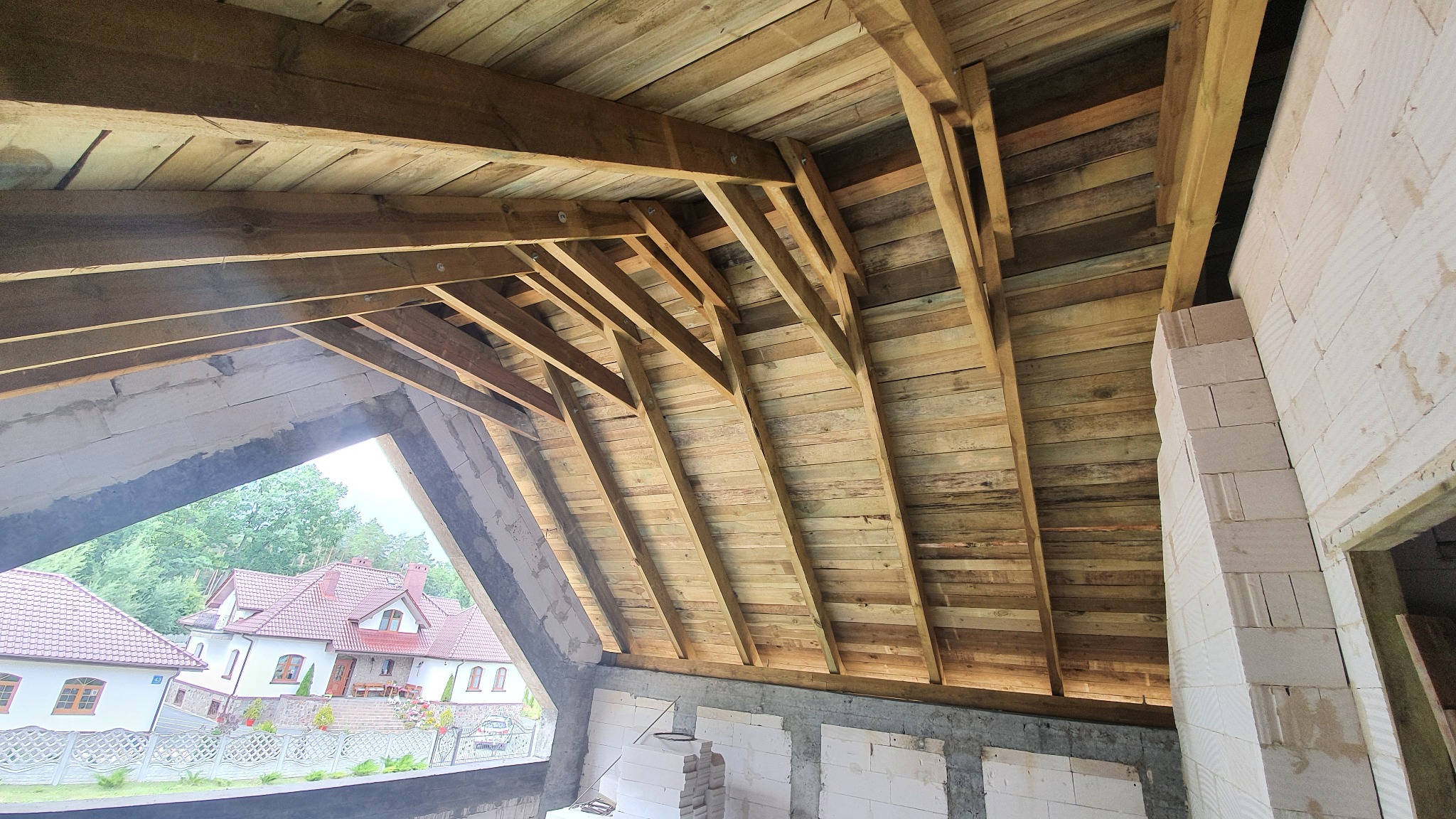 Wnętrze budowanego domu z widoczną drewnianą konstrukcją dachu i ścianami z białych bloczków, przez okno widać domy mieszkalne.