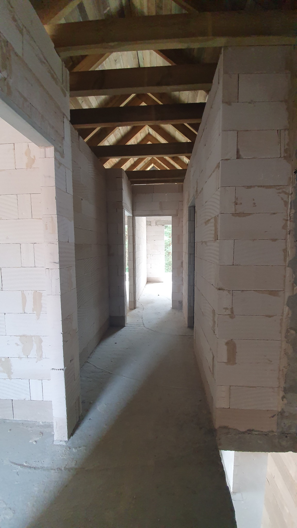 Wnętrze domu w trakcie budowy: widok na korytarz z surowymi ścianami z białych bloczków i drewnianą konstrukcją dachu, surowy beton na podłodze.