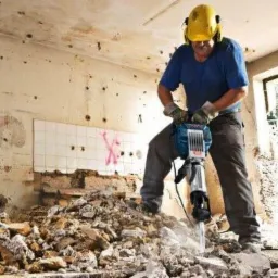 Pracownik w kasku używa młota pneumatycznego do wyburzania ściany w Warszawie, widoczne gruz i fragmenty płytek.