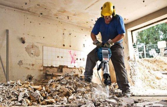 Pracownik w kasku używa młota pneumatycznego do wyburzania ściany w Warszawie, widoczne gruz i fragmenty płytek.
