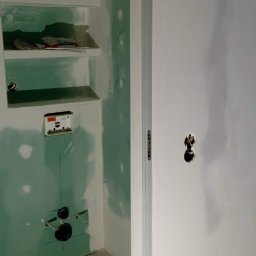 Rafal Witkowski - Wnętrze pomieszczenia w trakcie remontu: ściany pokryte płytą karton-gipsem, widoczne instalacje elektryczne i hydrauliczne, przygotowane półki wnękowe, świeżo wylana wylewka podłogowa.