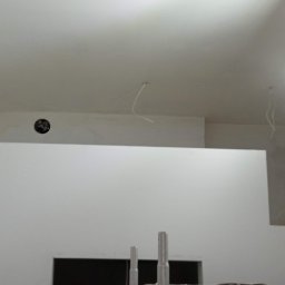 Rafal Witkowski - Wnętrze w trakcie prac wykończeniowych; ściany przygotowane do malowania lub położenia gładzi, widoczny żółty statyw z oświetleniem budowlanym, wystające kable i otwory w ścianach.