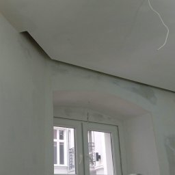 Rafal Witkowski - Wnętrze pokoju w trakcie remontu, widoczne ślady po gładzi na ścianach i suficie, zarys okna w tle oraz wystający kabel ze sufitu.