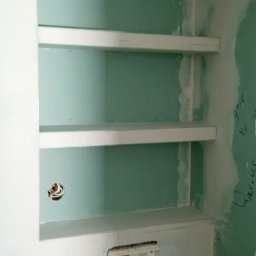 Rafal Witkowski - Niewykończona wnęka z trzema półkami w ścianie z płyt kartonowo-gipsowych, widoczne ślady szpachlowania i zarys instalacji elektrycznej w puszce.