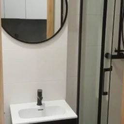 Nowoczesna łazienka z białą umywalką na czarnej szafce, okrągłym lustrem w czarnej ramie i kabiną prysznicową z czarnymi profilami.