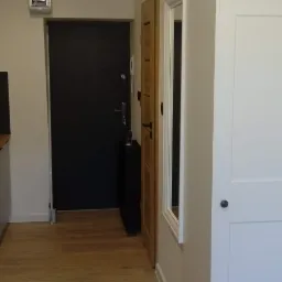 Wnętrze mieszkania z widoczną czarną, antywłamaniową  drzwi wejściowymi, drewnianymi drzwiami wewnętrznymi, białą szafą wnękową i panelami podłogowymi w odcieniach drewna.