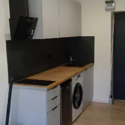 Nowoczesna kuchnia w mieszkaniu z szarymi szafkami, drewnianym blatem, czarnym panelem ściennym i pralką w zabudowie, widoczny kosz z narzędziami.