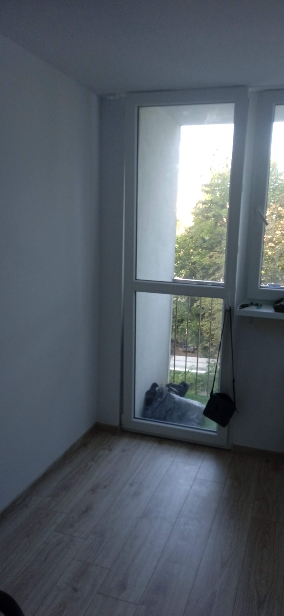 Wnętrze pokoju z świeżo pomalowanymi ścianami na biało, widok na balkon przez białe okno balkonowe, jasna drewniana podłoga.