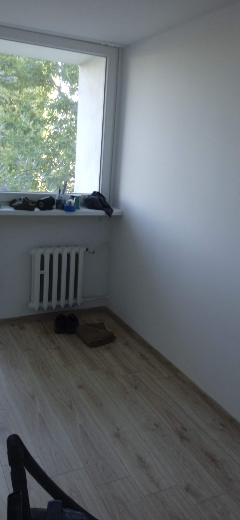 Pomalowane na biało wnętrze pokoju z drewnianą podłogą, grzejnikiem pod oknem i przedmiotami na parapecie.