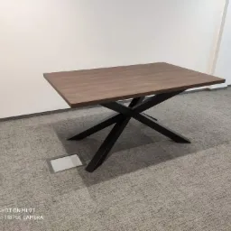 Stół z drewnianym blatem o ciemnobrązowym odcieniu i czarną, metalową podstawą w kształcie litery X, na tle białej ściany i szarej wykładziny podłogowej.