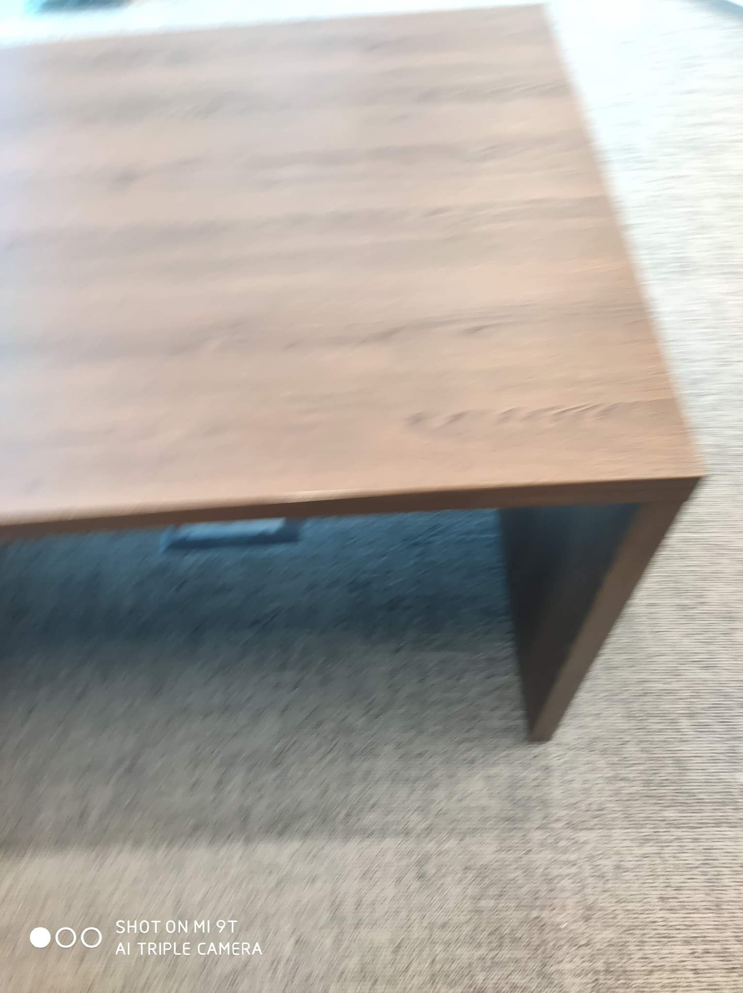 Fragment blatu z litego drewna o naturalnym odcieniu, widoczny z góry pod kątem, z wyraźną strukturą drewna i krawędzią. Niejasne tło sugeruje wykładzinę podłogową.