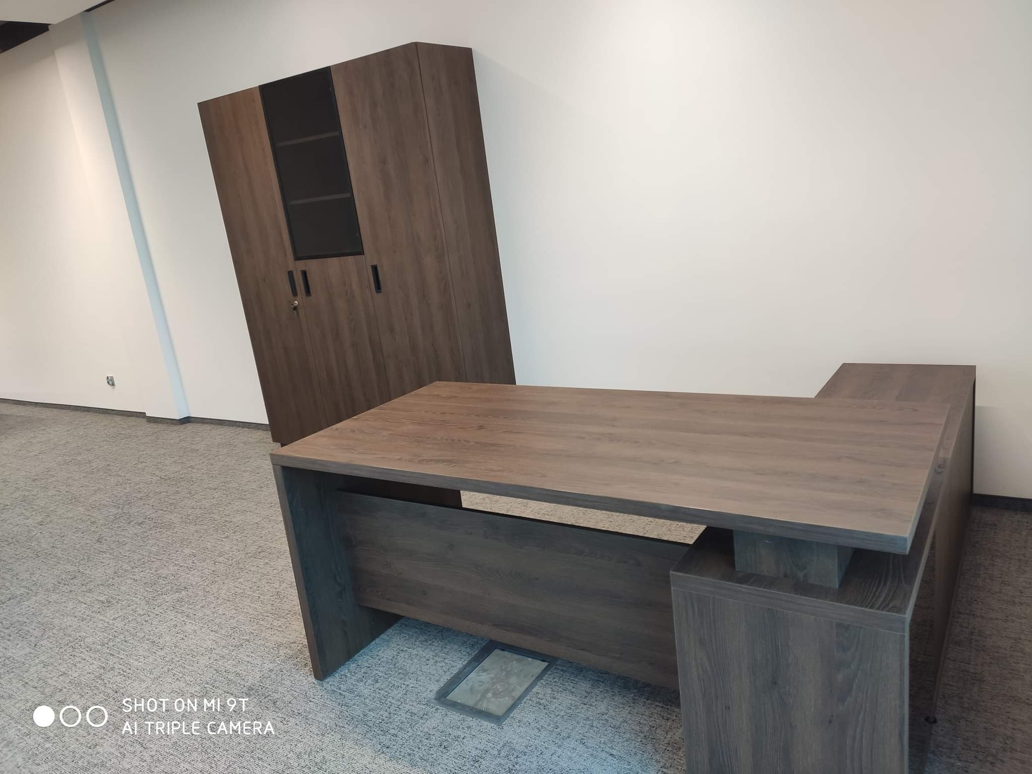 Biurko narożne z ciemnego drewna i szafa z przeszklonymi drzwiami w tle, widoczne na tle białej ściany i szarej wykładziny.