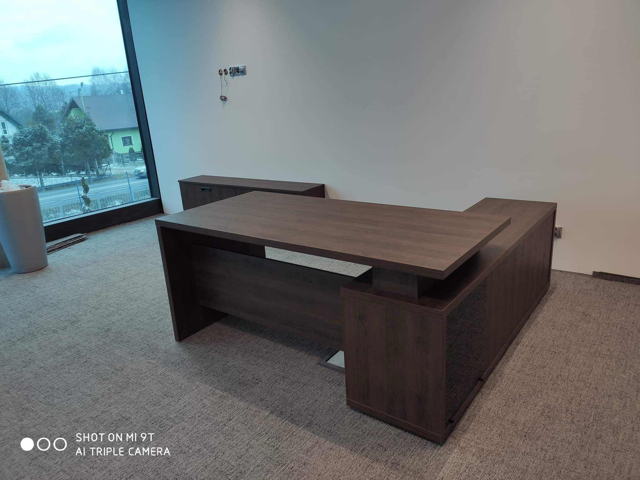 Nowoczesne biurko narożne o ciemnym, drewnopodobnym wykończeniu w przestronnym biurze z dużym oknem i widokiem na domy. Biurko z dodatkowym blatem i szafką w tle.