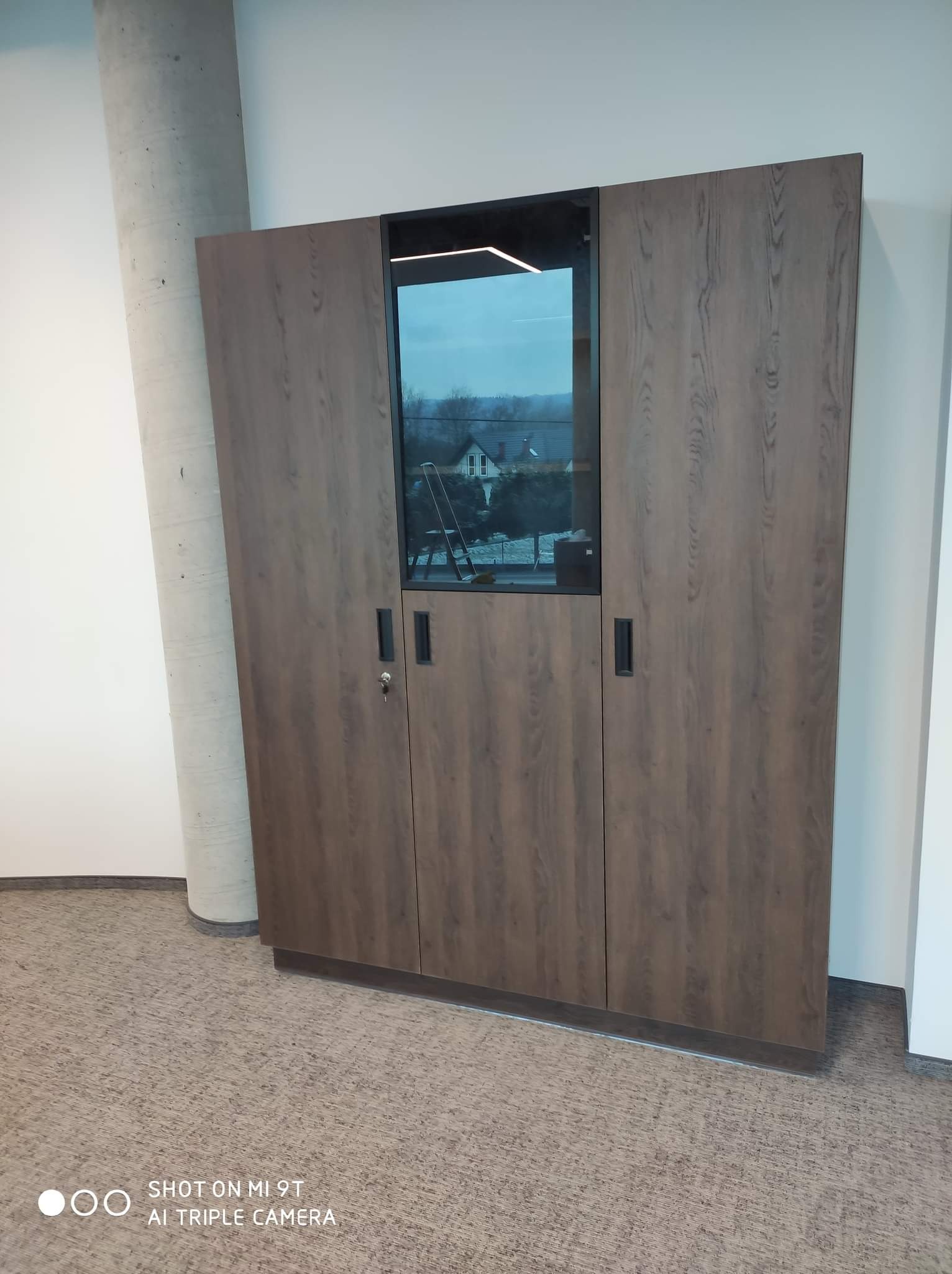 Wysoka, trzydrzwiowa szafa z okleiną drewnopodobną i przeszklonym środkowym panelem z widokiem na zewnątrz, zamek w dolnej części lewych drzwi, minimalistyczne uchwyty.