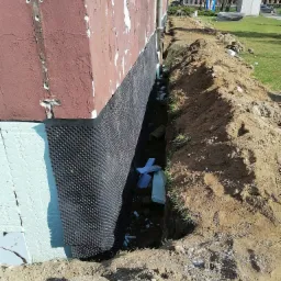 Odkopany fundament starego budynku z widoczną warstwą izolacji termicznej w kolorze jasnoniebieskim oraz folią kubełkową, zabezpieczony wykop z pryzmą ziemi, w tle budynek i trawnik.