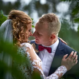 Para młoda w zbliżeniu, panna młoda w koronkowej sukni i welonie, pan młody w granatowym garniturze z bordową muchą, oboje patrzą sobie w oczy na tle zieleni.