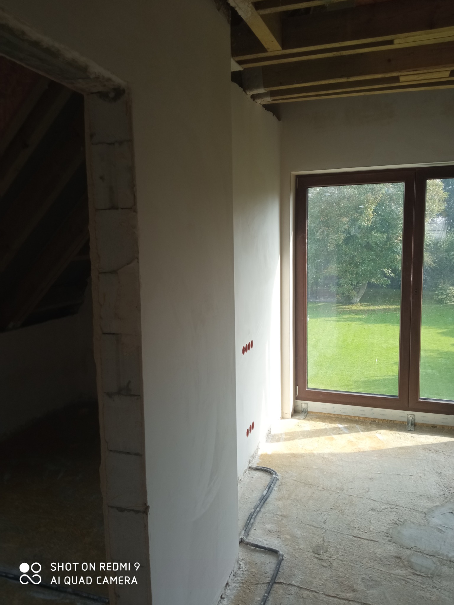 Wnętrze w trakcie tynkowania maszynowego: świeżo otynkowana ściana z widocznymi puszkami elektrycznymi, surowy strop drewniany i okno z widokiem na zielony ogród.