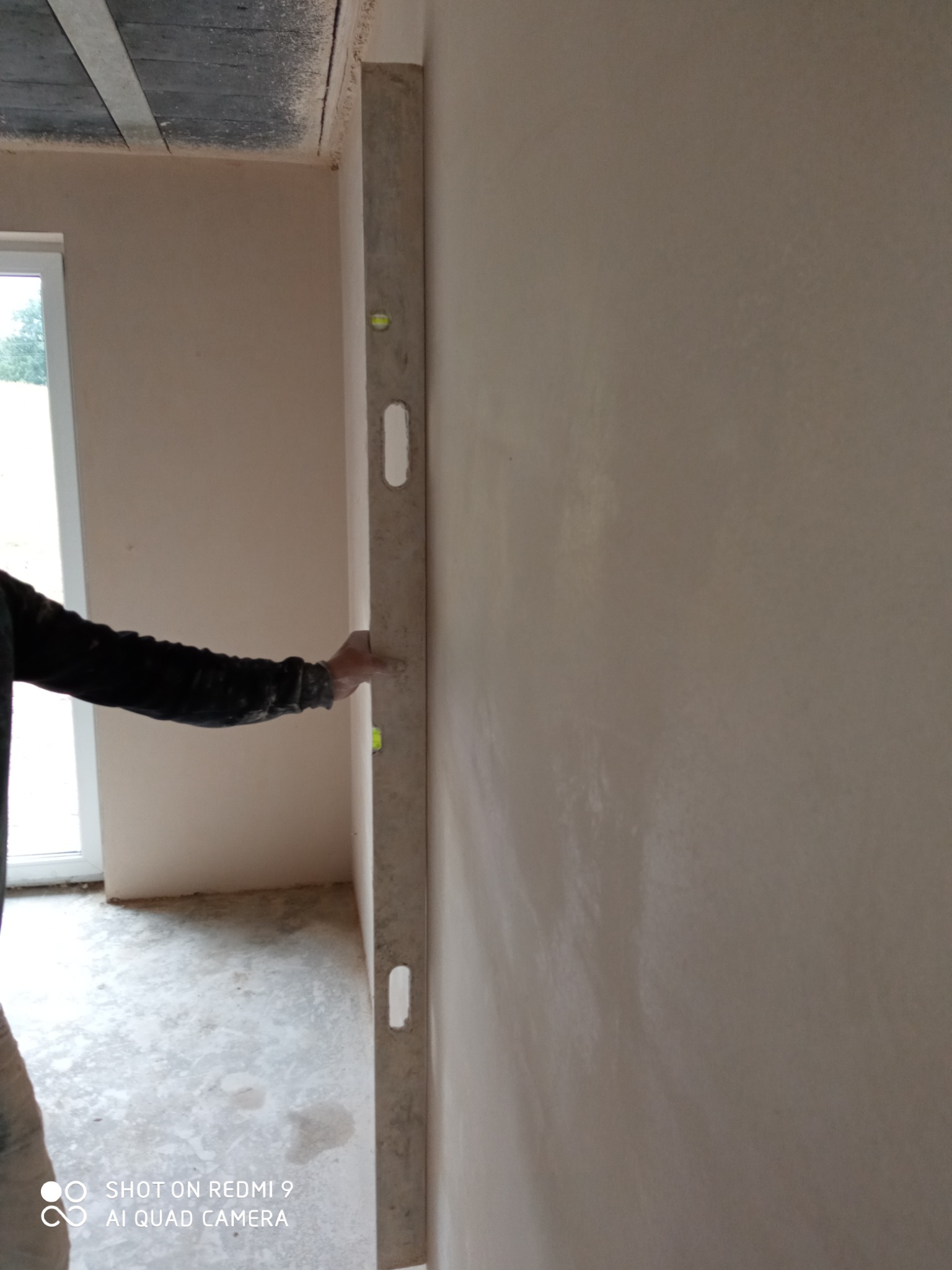 Sprawdzanie pionu ściany po nałożeniu tynku maszynowego za pomocą poziomicy. W tle widoczne okno i surowy sufit.