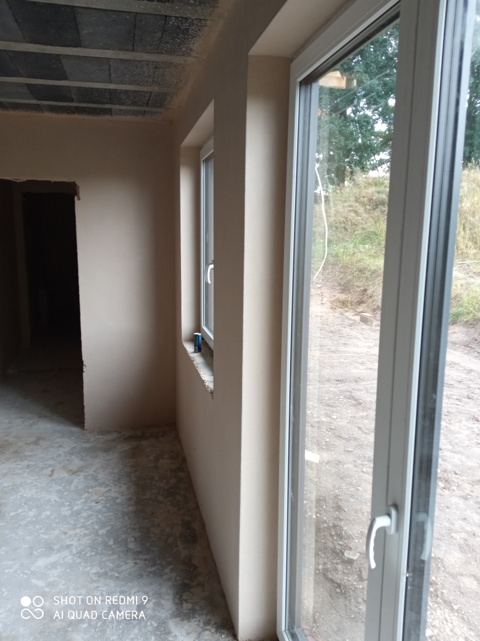 Wnętrze w stanie surowym po nałożeniu gładzi gipsowej, widoczne okno PCV z widokiem na zewnątrz i sufit z betonowych płyt stropowych.